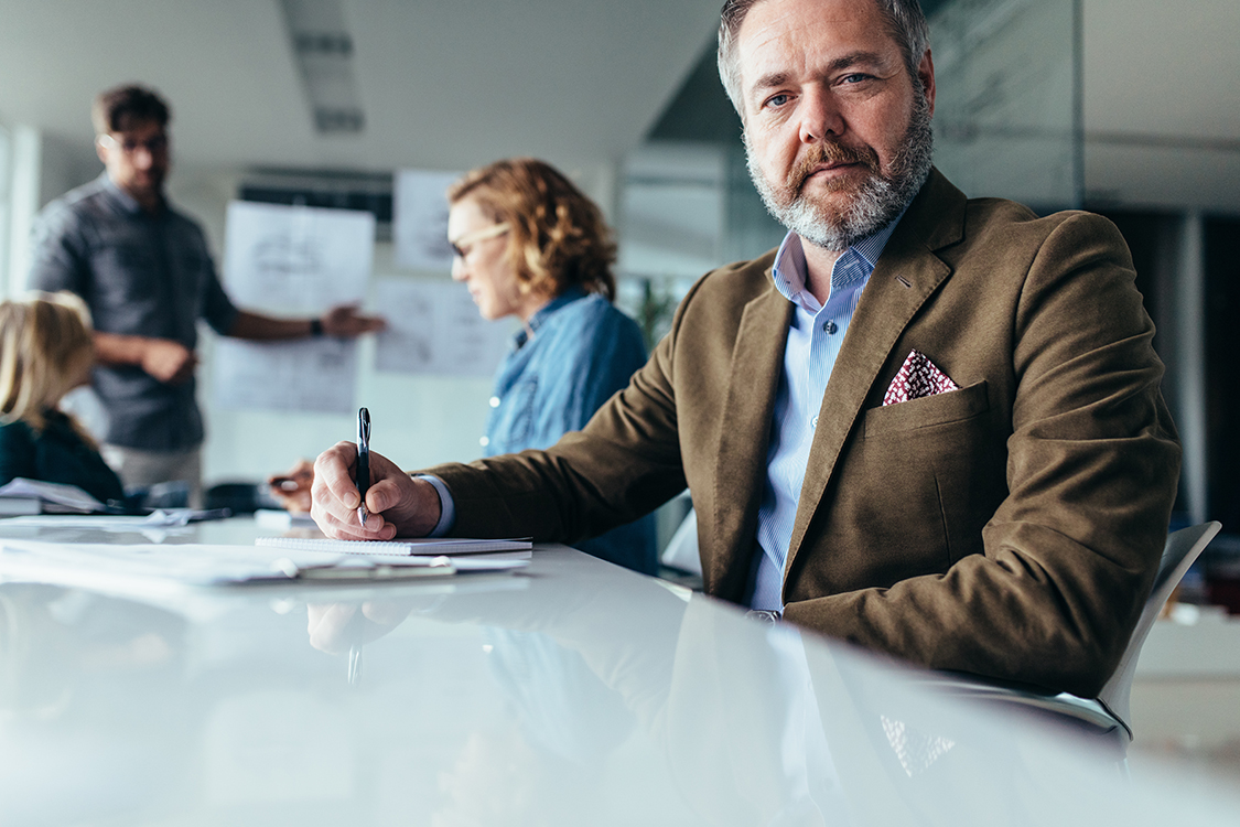 Mature businessman sitting in conference room with colleagues in background. Senior man sitting in board room during presentation.
