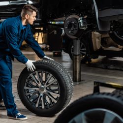 young mechanic holding car wheel near raised car in workshop