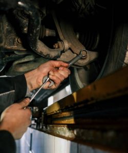Mechanic's hand holding a wrench, working underneath a car on a lift. The view shows a close-up of the suspension and exhaust system in a garage setting.